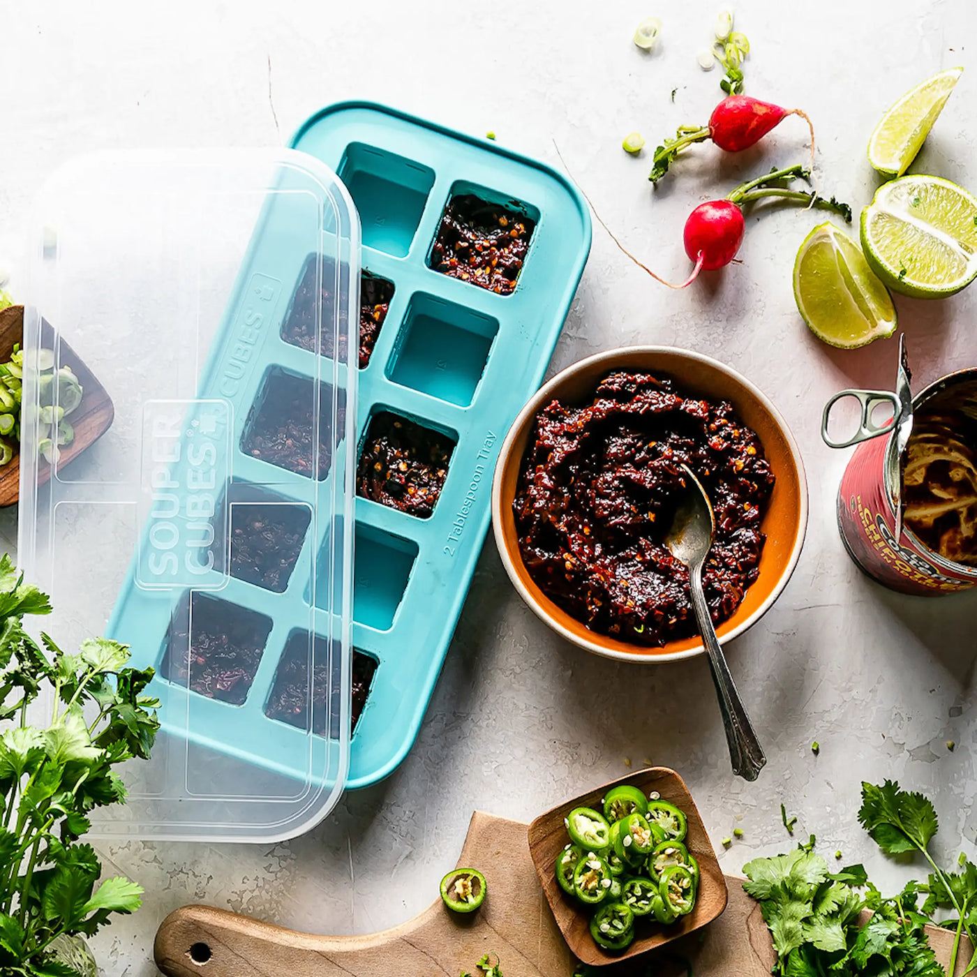 Turkos Souper Cubes-form fylld med mat i portionsstorlekar, omgiven av en skål med röd sås, limeklyftor, rädisor, koriander och skivade gröna chilifrukter på en ljus bänkskiva.