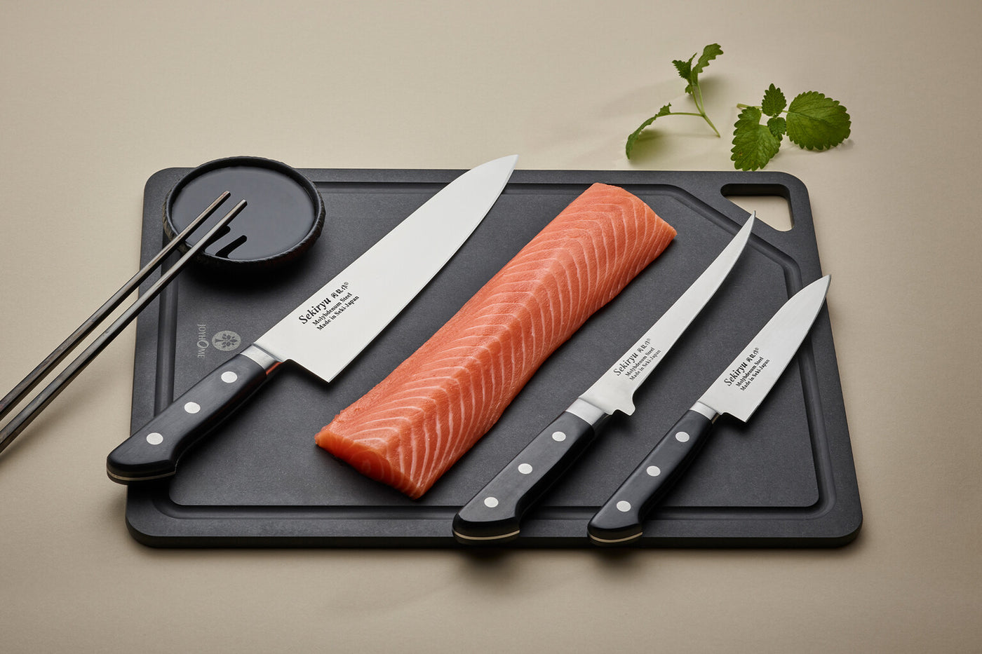 Tre japanska knivar från Sekiryu ligger på en svart skärbräda tillsammans med en bit färsk lax, ett fat med soja och ett par ätpinnar.
