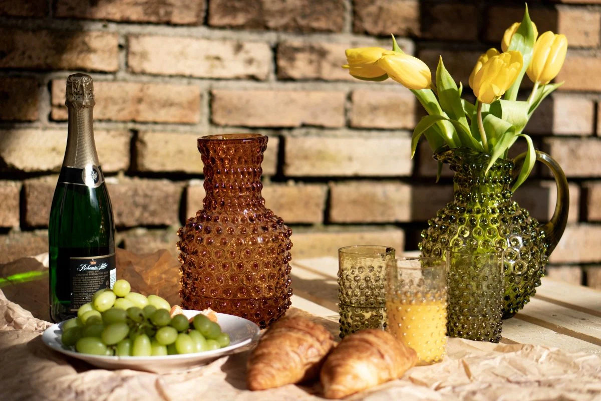 Hobnail-karaffer och glas från Klimchi i bärnsten och olivgrönt – handgjorda glasprodukter i bubblig design, perfekta för stilfull utomhusdukning med croissanter och juice.